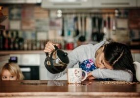 Tired mother, trying to pour coffee in the morning. Woman lying on kitchen table after sleepless night, trying to drink coffee