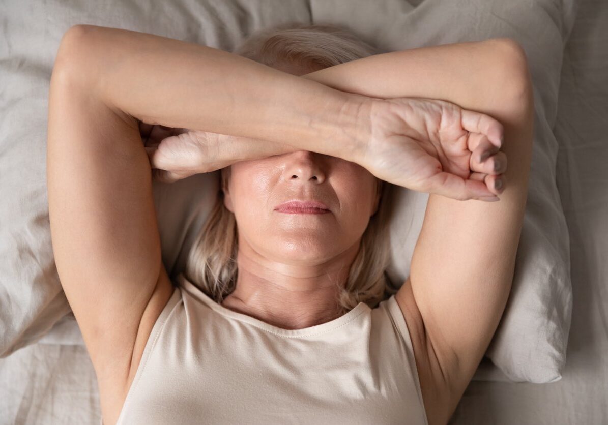 Sleepless mature woman suffering from insomnia close up, lying in bed, older female covering eyes with hands, trying to sleep, nightmares or depression, feeling headache or migraine